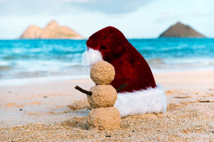 A snowman made of sand positioned next to a Christmas hat on a Hawaiian beach
